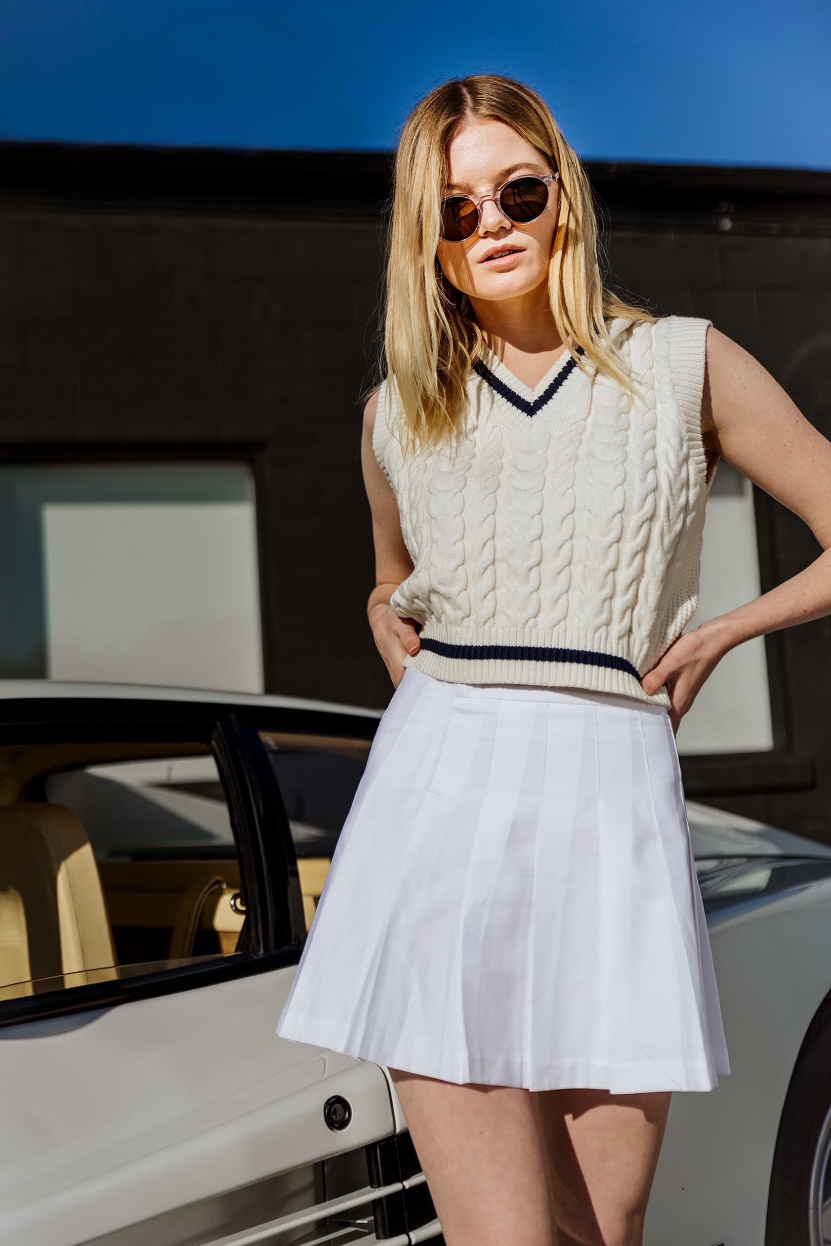 A model wearing Waid sunglasses and a white knit vest beside a Ferrari.