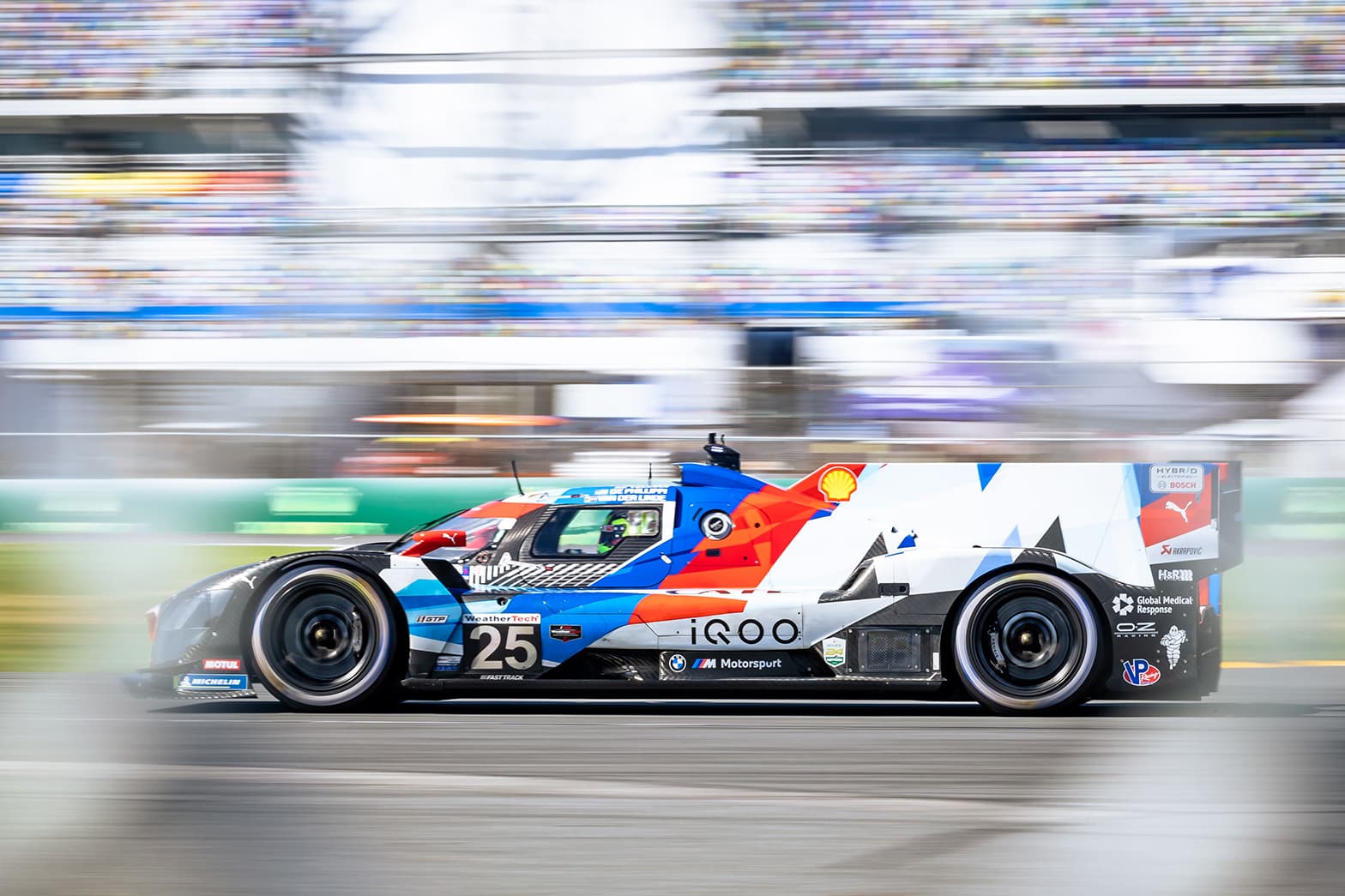 A BMW prototype racing past blurred grandstands in daylight.