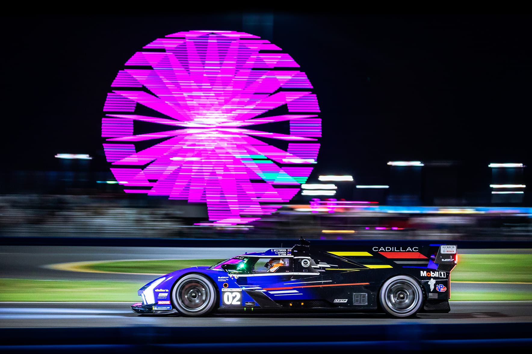 A Cadillac prototype racing beneath a pink-lit Ferris wheel.