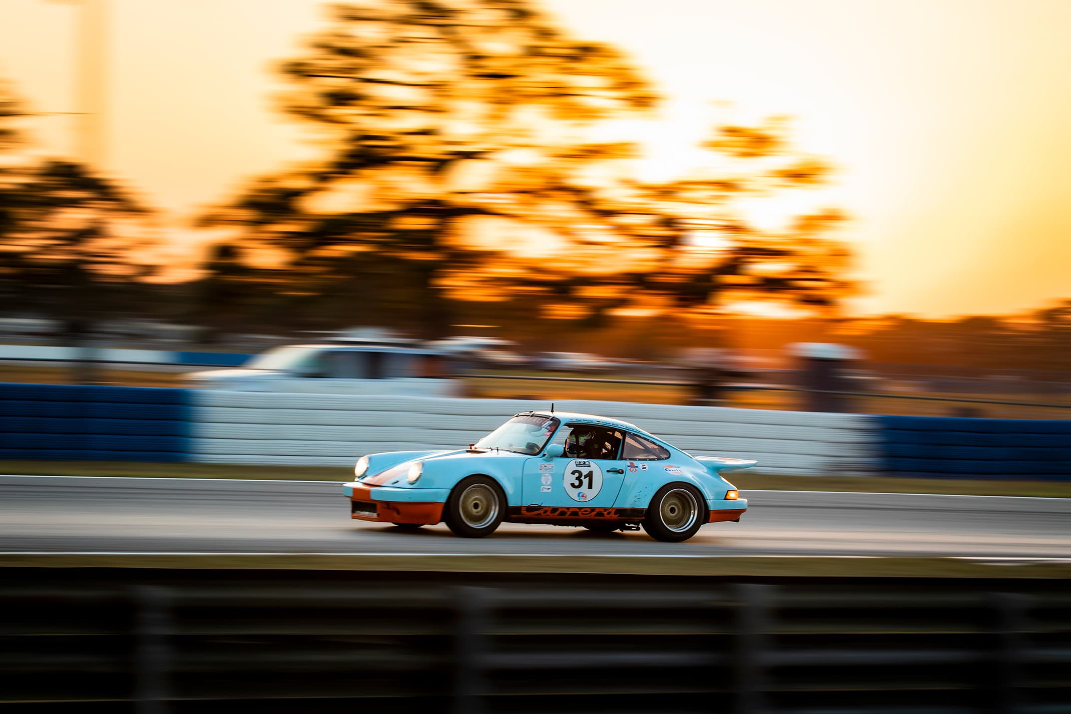 A light blue and orange number 31 Porsche Carrera racing past at sunset.