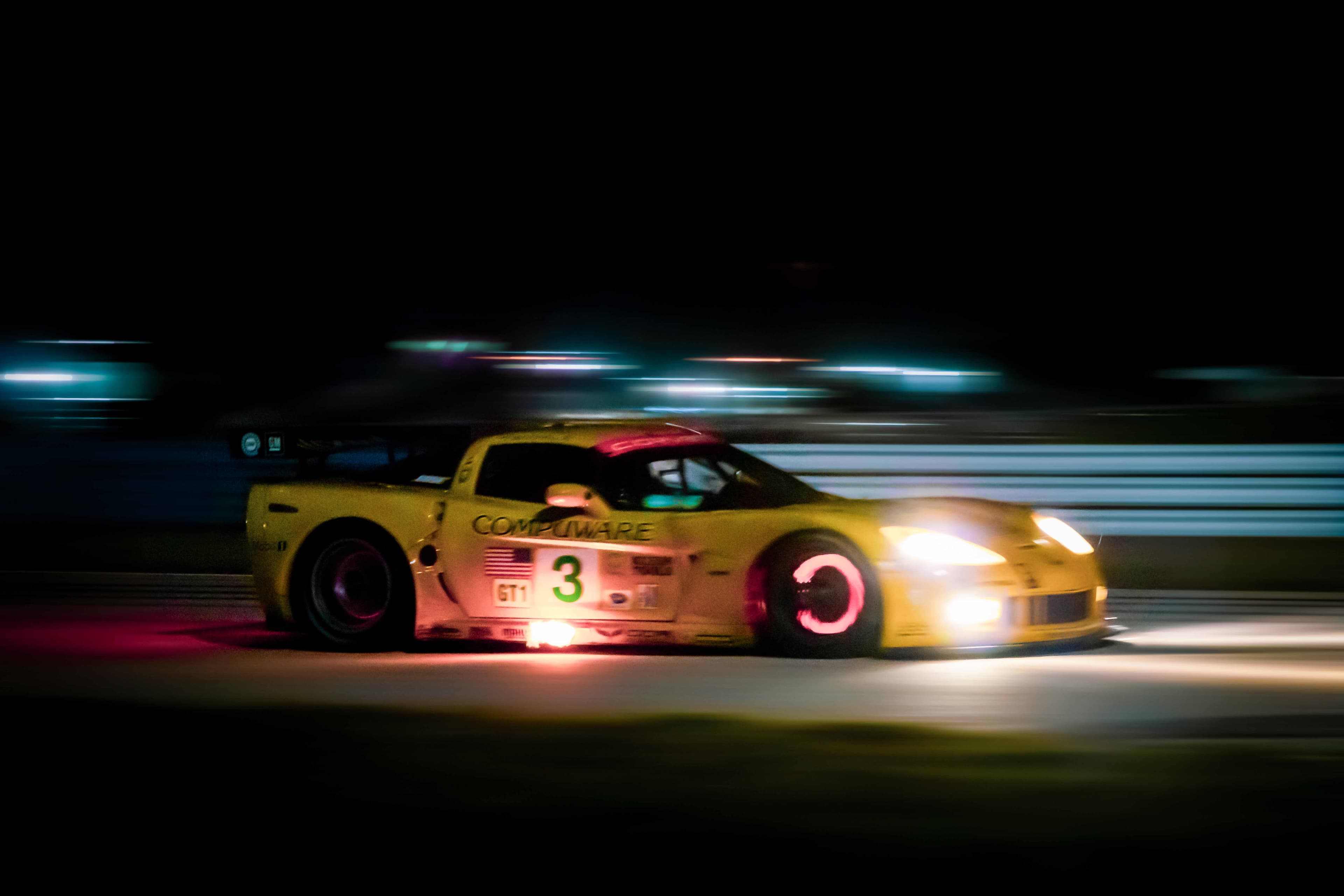 A yellow number 3 Corvette racing at night with glowing brakes and flames beneath its side exhaust.