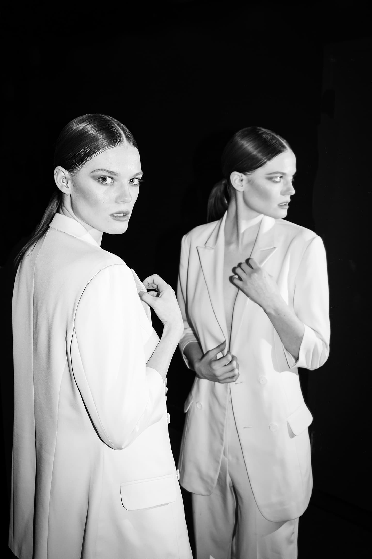 Black-and-white portrait of a model in a white suit beside her reflection.