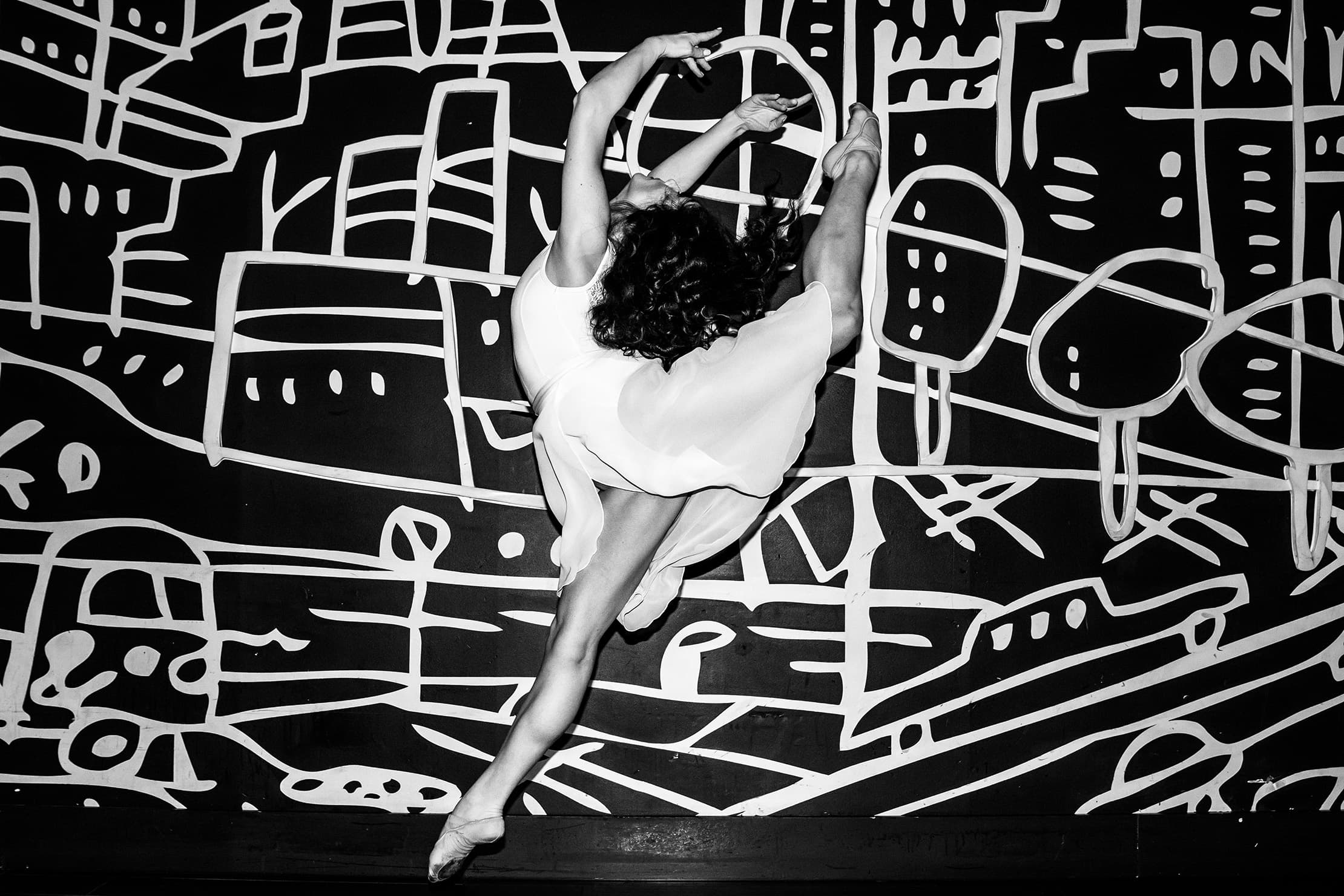 Black-and-white photograph of a dancer leaping in front of a bold abstract mural.