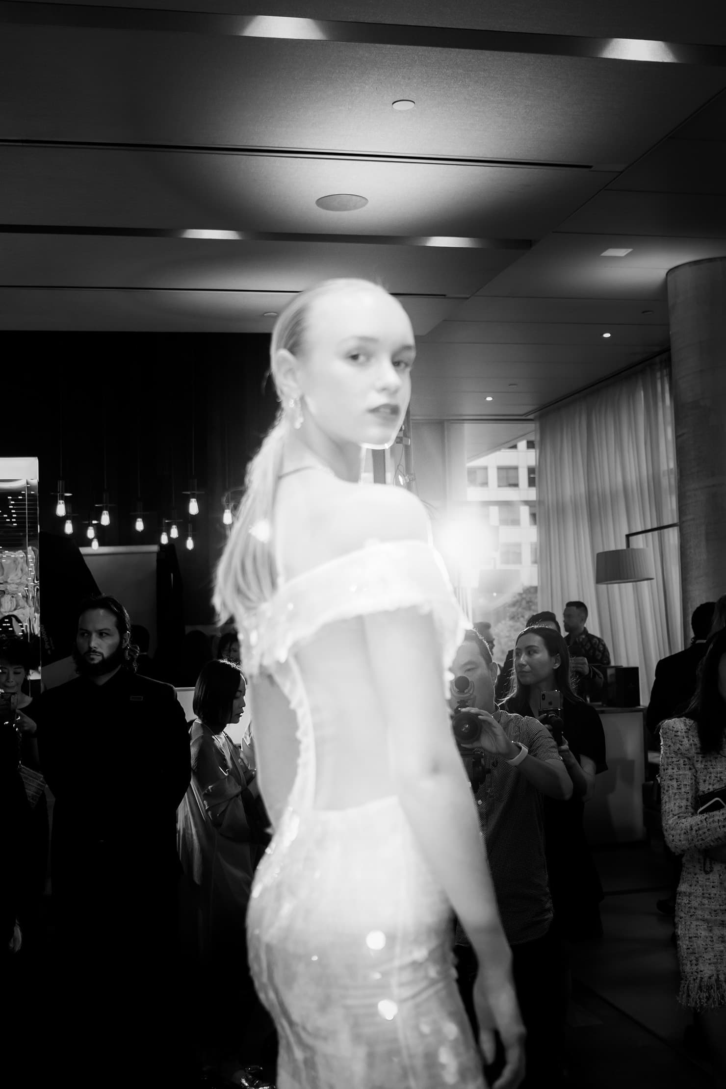 Black-and-white photograph of a runway model passing an audience with bright light behind her.