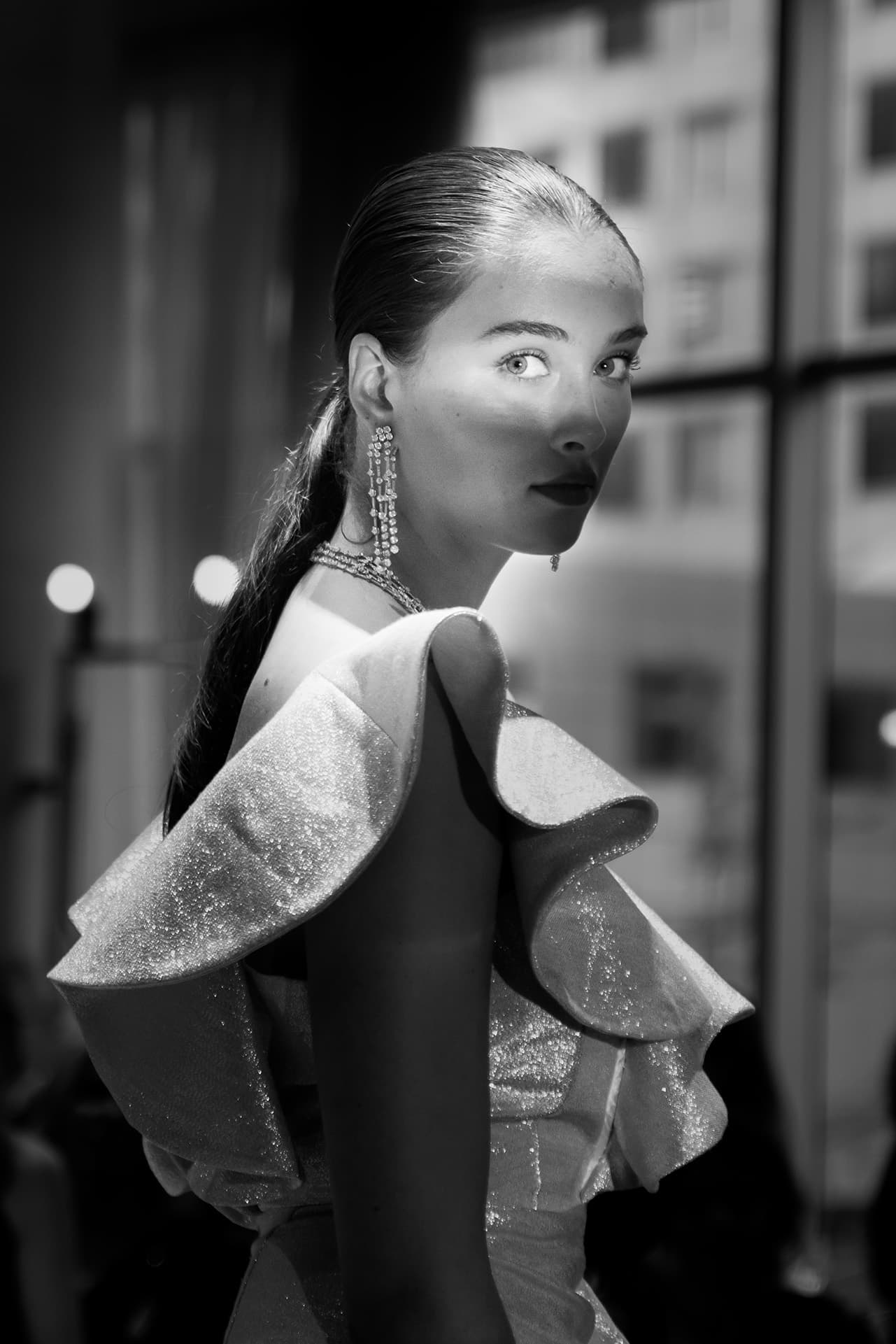 Black-and-white portrait of a runway model in a sculptural ruffled dress beside a window.