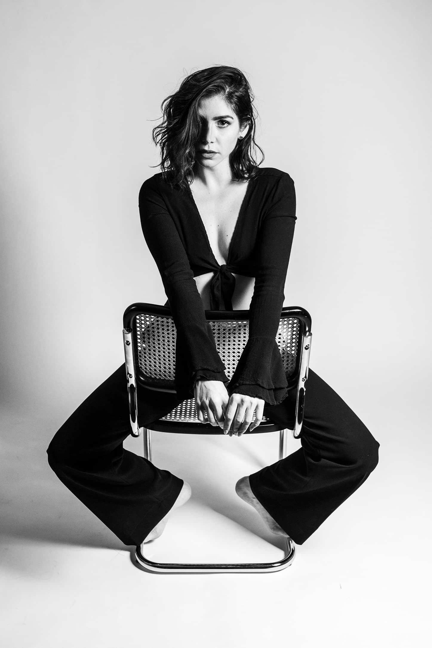Black-and-white portrait of a model seated backwards on a woven chair.