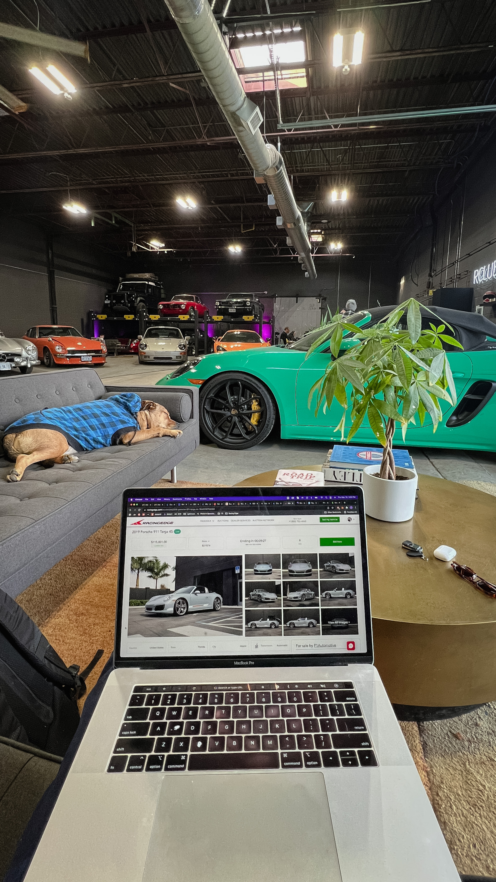 A laptop displaying a RacingEdge Porsche auction in an automotive clubhouse, with sports cars and a dog resting on a sofa.